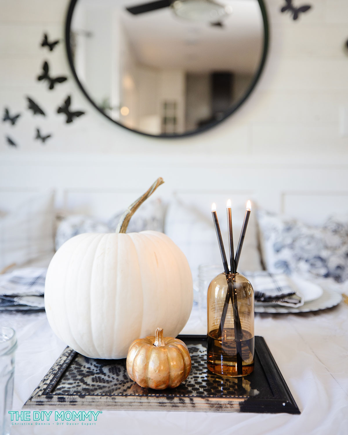 A larger faux white pumpkin with a smaller gold pumpkin as a table centerpiece along with 3 candles in a single vase. 
