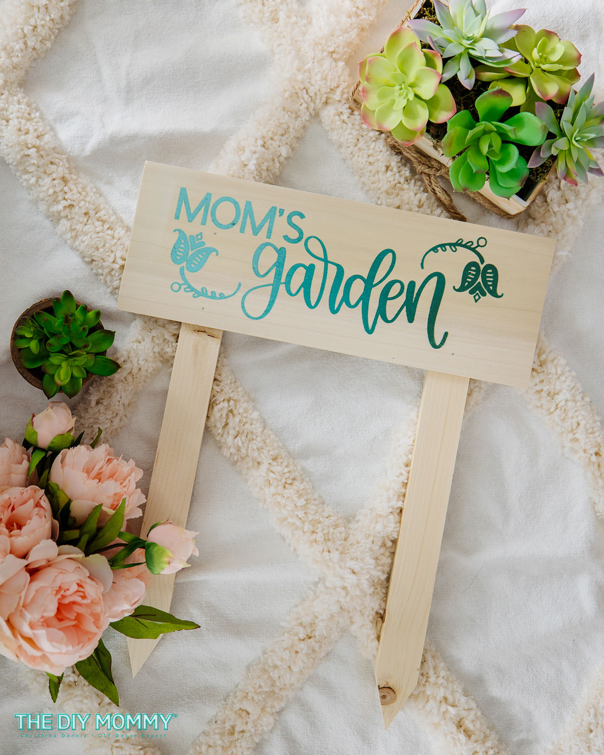 A wooden sign with Mom's Garden in green and teal writing is displayed with flowers.