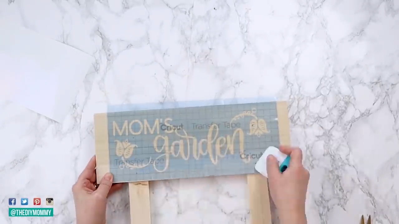 A stencil, "mom's garden", is being applied with transfer tape to a wooden sign. 